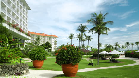 Gardener-At-The-Hotel-Busy-Mowing-The-Lawn-On-A-Sunny-Day-With-Scenery-Of-Palm-Trees-And-Potted-Floral-Plants