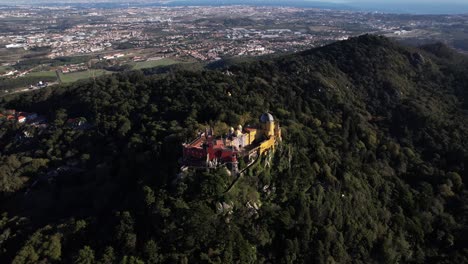 Drone-shot-of-historic-Pena-Palace-castle-illuminated-by-sunlight-standing-a-top-of-a-hill-in-Sintra-mountains-above-town-of-Sintra,-Portugal