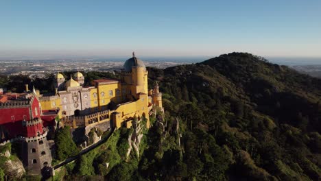Aerial-revealing-shot-of-historic-Pena-Palace-castle-exterior-architectural-design-illuminated-with-sunlight-standing-a-top-of-a-hill-in-Sintra-mountains-above-town-of-Sintra,-Portugal
