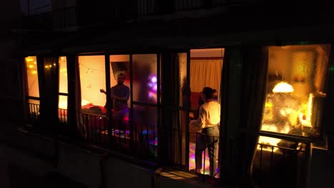 Drone-shot-from-outside-of-a-group-of-people-celebrating-and-dancing-in-an-apartment-at-night,-multicolored-lights