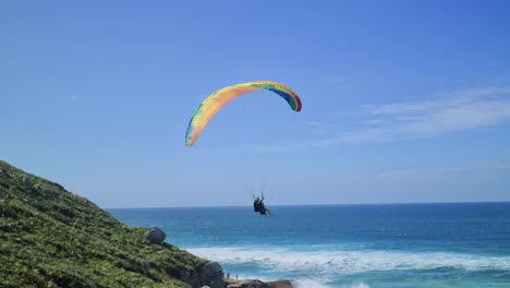 Paragliding-flight-scene-in-front-of-the-beach-with-rocks-mountains-and-sand-with-people-having-fun-in-the-flight-with-sea-waves-air-sport