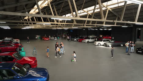 Fans-admiring-vehicles-in-retro-BMW-e30-car-club-warehouse-meeting,-aerial-view-reversing