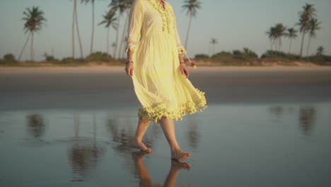 Beautiful-Indian-woman-wearing-a-yellow-summer-dress-walking-on-a-tropical-beach-shoreline,-with-palm-trees-in-the-background