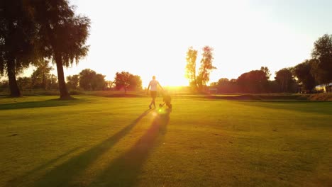 Low-Winkel-Drohne-Schoss-Nach-Einem-Golfer,-Der-Während-Des-Atemberaubenden-Sonnenuntergangs-Mit-Einem-Trolley-Auf-Dem-Golfplatz-Spazieren-Ging