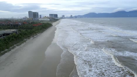 Beautiful-aerial-shot-of-Da-Nang,-An-Bang-Beach,-Vietnam-with-the-waves-rolling-on-to-the-beach-and-skyscrapers-in-the-background