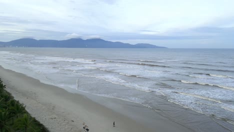 Rotation-aerial-shot-of-waves-hitting-a-beach-on-a-cloudy-day-and-mountains-in-the-background
