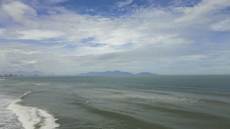 Waves-in-the-ocean-breaking-towards-the-coast-with-big-fluffy-clouds-in-the-sky-and-mountains-in-the-horizon