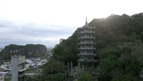 Impresionante-Antena-De-Un-Templo-En-Las-Montañas-Verdes-Asiáticas-Que-Se-Elevan-Sobre-Una-Ciudad-Durante-La-Puesta-De-Sol