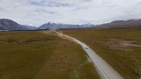 oncoming-car-on-alpine-dirt-road-between-dry-plains-and-snow-capped-mountain-range-in-background