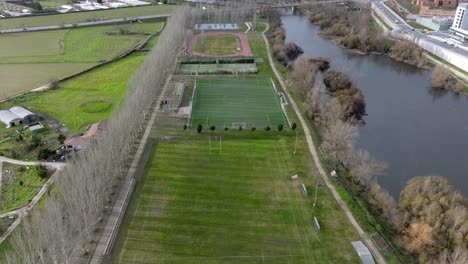 Fliegen-über-Grüne-Fußballfelder-Und-Laufplätze-In-Der-Nähe-Des-Blauen-Flusses-In-Spanien