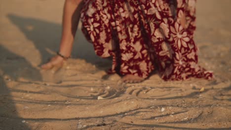 Beautiful-Indian-woman-wearing-a-red-summer-dress,-moving-sand-with-her-hands,-on-a-tropical-beach