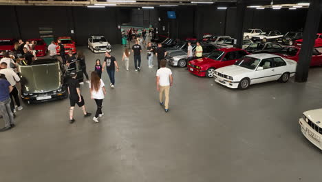 Fans-walking-around-BMW-e30-car-club-meeting-admiring-vehicles,-aerial-view-following