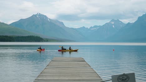 Family-of-3-paddling-kayaks-on-lake-in-glacier-national-park-in-slow-motion-with-beautiful-mountain-backdrop-and-boat-dock-in-the-foreground