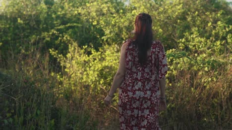 Out-of-focus-brunette-Indian-woman-walking-away-through-high-grass-and-trees,-wearing-a-red-summer-dress