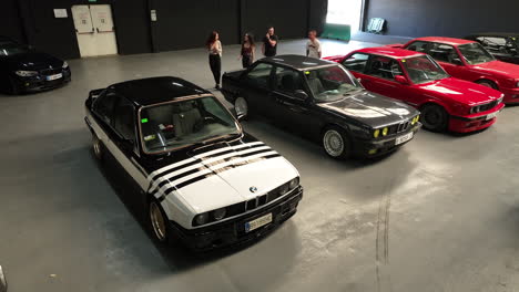 Custom-BMW-e30-vehicles-parked-in-old-school-classic-warehouse-car-show,-Aerial-slow-orbit-view