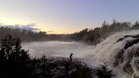 People-walking-on-slippery-rocks-next-to-a-huge-and-violent-waterfall-during-sunset-in-the-forest
