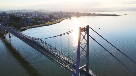 Drone-aerial-scene-of-Hercílio-Luz-bridge-at-dusk-at-sunset-with-large-capital-city-urban-complex-Florianópolis-late-afternoon-with-metallic-suspension-bridge-sun-setting-with-bridge