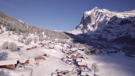 Empujando-Sobre-El-Pueblo-De-Montaña-Cubierto-De-Nieve-De-Grindelwald-En-Dirección-Al-Monte-Wetterhorn-En-Un-Día-De-Invierno-De-Pájaro-Azul