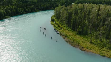 Aerial-drone-orbit-over-large-group-of-King-Alaskan-salmon-fisherman,-wading-and-fishing-in-pristine-blue-water-on-lush-green-riverbed-shoreline