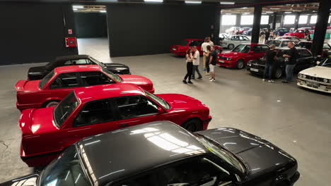 Fans-hanging-out-around-colourful-BMW-e30-vehicles-parked-in-warehouse-club-meeting-event,-orbiting-aerial-view