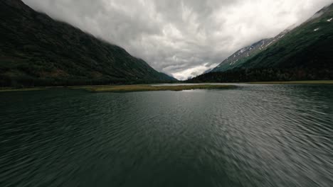 FPV-aerial-drone-flying-over-Alaskan-lake-surrounded-by-mountains-and-lush-flora-on-cloudy-day