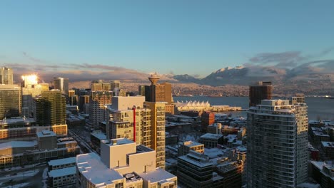 Vancouver-Edificios-Comerciales-Cubiertos-De-Nieve-Durante-La-Puesta-De-Sol-Con-Montañas-Cubiertas-De-Nieve-En-El-Fondo