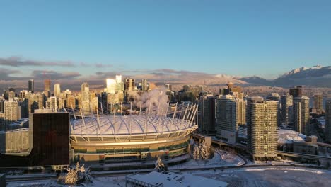 Vista-Aérea-De-Los-Edificios-Comerciales-De-Vancouver-Alrededor-Del-Estadio-Bc-Place-Con-Montañas-Cubiertas-De-Nieve-En-El-Fondo