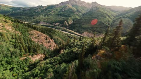 FPV-aerial-drone-flying-over-Alaskan-lush-green-forest-mountains-and-rocky-canyons-and-under-old-metal-train-bridge-near-Denali-National-Park-with-sun-flares