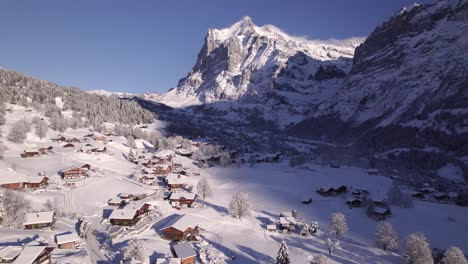 pushing-in-over-snowy-mountain-village-of-Grindelwald-on-a-sunny-winter-day,-flying-towards-Mount-Wetterhorn-in-Switzerland