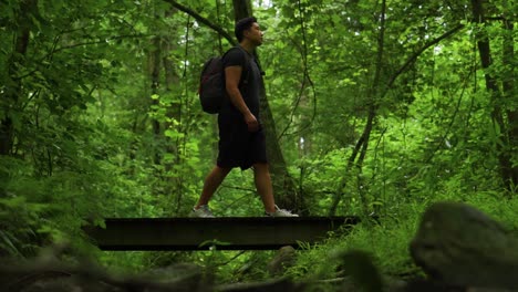 Young-man-walks-over-a-foot-bridge-during-a-hike-in-the-forest-carrying-a-backpack-and-gazing-up-at-the-green-trees