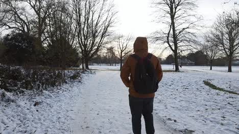 Man-With-Backpack-Walking-In-The-Snowy-Path-To-Brooklands-Lake-During-Winter-In-Dartford,-England,-UK