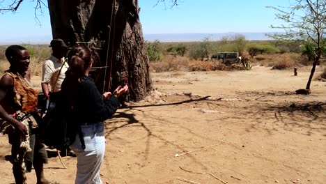Mujer-Turista-Aprendiendo-A-Disparar-Con-Arco-Sin-éxito-Con-La-Gente-De-La-Tribu-Hadzabe-En-Tanzania