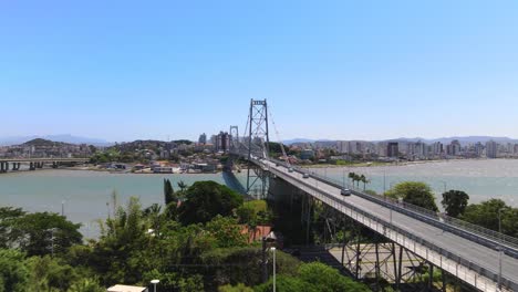 Drone-aerial-scene-of-Hercílio-Luz-bridge-with-large-capital-city-urban-complex-Florianópolis-with-metallic-suspension-bridge-sun-setting-with-bridge-and-urbanism-center