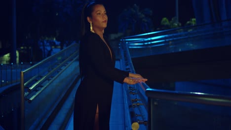 A-woman-in-a-black-dress-leans-on-the-railing-of-a-walkway-in-the-city-at-night