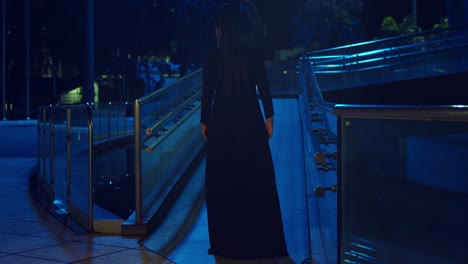 Mujer-Hispana-Camina-Por-Una-Pasarela-En-La-Ciudad-Con-Un-Vestido-Negro-Y-Luces-Azules-En-El-Fondo