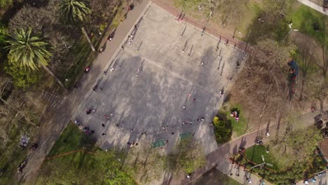 Group-of-kids-skating-in-skate-park-during-sunny-day-surrounded-by-palm-trees-in-Buenos-Aires---Aerial-top-down-circling-shot