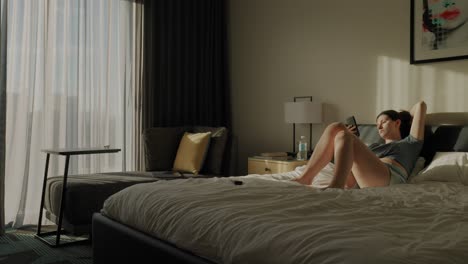 Woman-laying-in-bed-in-the-morning-scrolling-on-her-phone-|-Wide-shot