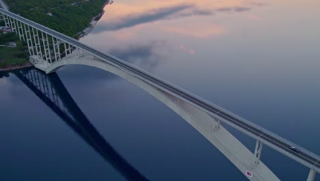 Krk-arch-concrete-bridge-crossing-water-during-sunrise,-aerial