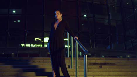 Young-woman-stands-on-the-stairway-of-a-glass-building-in-the-city-at-night-in-a-black-dress