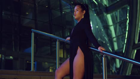 Young-woman-in-a-black-dress-stands-on-the-stairway-of-a-glass-building-in-the-city-at-night