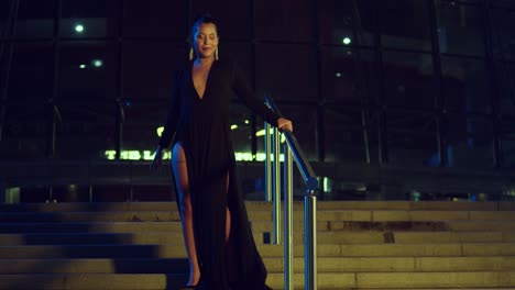 Sexy-lady-in-a-black-dress-walking-down-a-stairway-of-a-glass-building-at-night-in-the-city