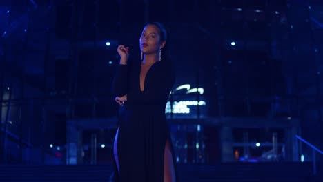 Young-hispanic-woman-stands-in-the-city-in-a-black-dress-with-blue-lights-in-the-background
