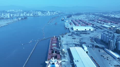 grain-loading-on-freighter-from-distribution-port-canal-in-Surrey-BC-Canada-top-birds-eye-view-overlooking-commercial-dock-as-other-cargo-ship-are-getting-loaded-with-wood-vehicles-drone-flyover-1-2