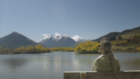 Blond-female-tourist-sitting-on-wooden-bench-enjoying-sunny-view-of-Glenorchy-lagoon