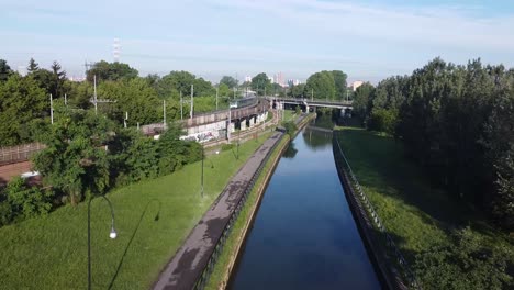 Passenger-train-riding-on-track-curve-in-Milan-city-near-water-canal,-aerial-view