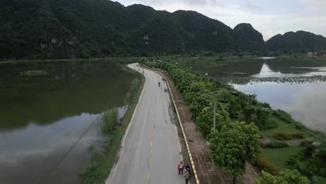 Cool-aerial-shot-following-people-on-bikes-on-a-asian-road-sounded-my-water-and-stunning-green-mountains-in-Nin-Bihn,-Vietnam