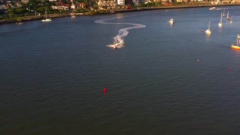Aerial-panning-view,-following-a-jetski-speeding-on-the-river-from-Hamilton-towards-Newstead-capturing-northern-riverside-residential-complex-view-at-sunset-golden-hour,-Brisbane-city,-Queensland