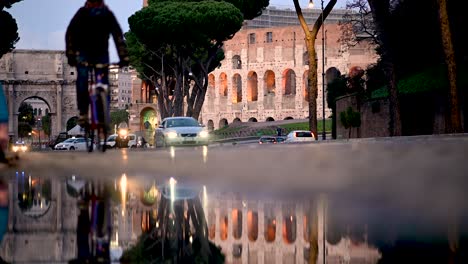 Cars,-bicycles-and-scooters-driving-past-reflection-in-puddle-outside-the-Colosseum,-Rome-Italy