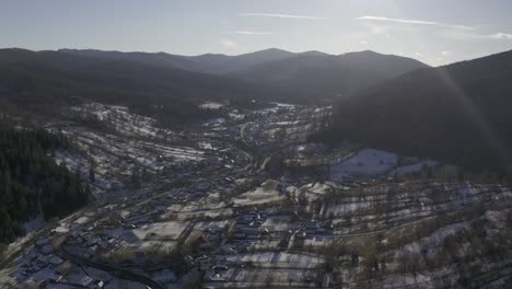 Drone-view-of-a-city-in-the-valley-below-a-mountain-range-lined-with-pine-trees-and-snow