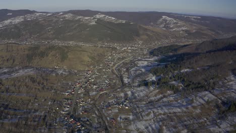 Drone-view-of-a-village-in-the-valley-below-a-mountain-range-lined-with-pine-trees-and-snow
